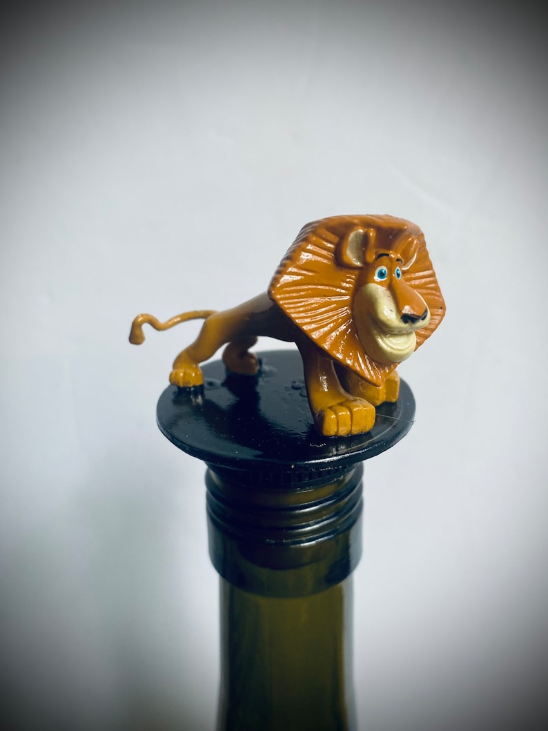 May include: A brown lion figurine with a blue eye and a black nose is sitting on top of a black bottle stopper. The stopper is on top of a green glass bottle.