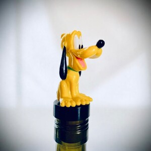 May include: A yellow and black plastic bottle stopper shaped like Pluto, a cartoon dog from Disney. The stopper is sitting on top of a wine bottle.