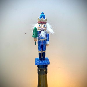 Op de afbeelding: Blauwe glitter-notenkraker flessenstop met een groene kerstboom in de hand, die een gouden kroon en een witte baard draagt.