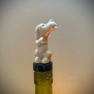 Puede incluir: Un tapón de botella de cerámica blanca con forma de perro sobre una botella de vidrio verde. La figura del perro tiene un acabado brillante con ojos negros y un tapón negro. La botella es parcialmente visible.