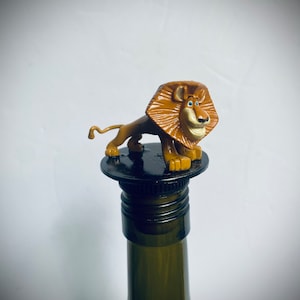 May include: A brown lion figurine with blue eyes on top of a black bottle stopper. The lion is standing on its hind legs with its front paws raised. The bottle stopper is on top of a green wine bottle.