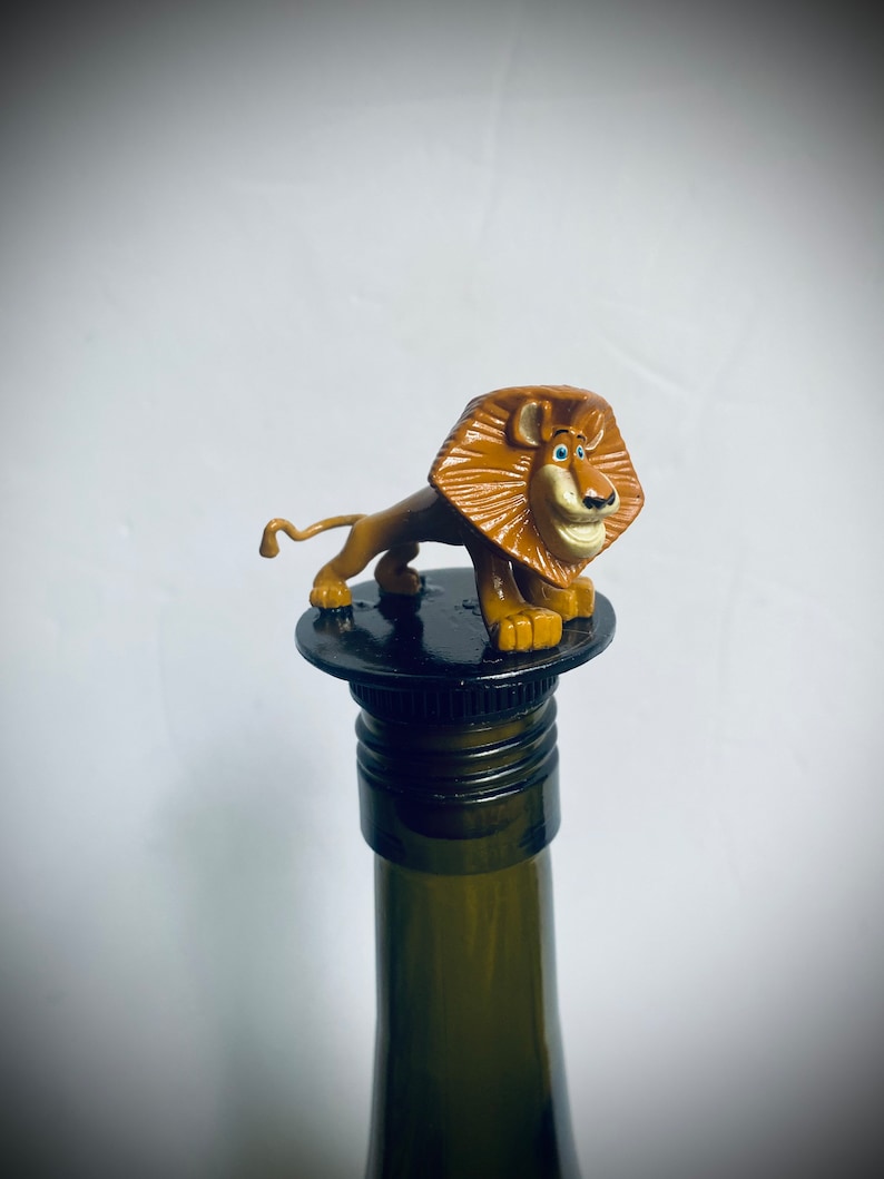 May include: A brown lion figurine with blue eyes on top of a black bottle stopper. The lion is standing on its hind legs with its front paws raised. The bottle stopper is on top of a green wine bottle.