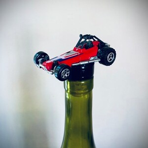May include: A red toy dune buggy with black wheels is used as a bottle stopper on a green glass wine bottle.