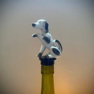 Pode incluir: Um tampão de garrafa de cerâmica branco e preto em forma de cão com uma base preta. O tampão está em cima de uma garrafa de vinho de vidro verde.
