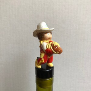 May include: A cowboy figurine with a white hat, brown hair, and red and yellow clothing riding a brown horse. The figurine is a bottle stopper.