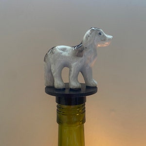 Puede incluir: Un tapón de botella de cerámica blanca en forma de perro con marcas negras. El tapón está sobre una base negra y una botella de vino verde.