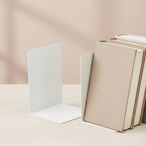 Puede incluir: Dos sujetalibros de metal blanco en forma de L con un acabado mate liso. Un sujetalibros se encuentra solo a la izquierda, mientras que el otro sostiene una pequeña pila de tres libros de tapa dura con cubiertas en tonos beige y crema.