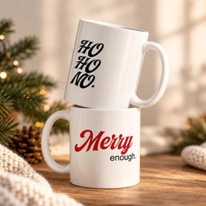 Könnte beinhalten: Zwei weiße Keramiktassen, die vertikal gestapelt sind. Die obere Tasse zeigt den schwarzen Text HO HO NO. Die untere Tasse zeigt den roten Text Merry und den schwarzen Text enough. Beide Tassen haben einen Standardhenkel und eine glatte Oberfläche.
