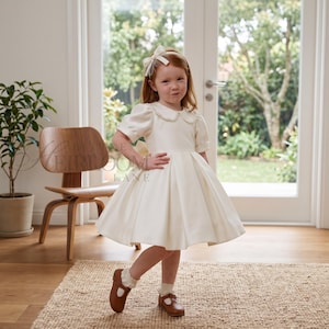 Puede incluir: Un vestido color crema con mangas cortas abullonadas, cuello redondo y falda plisada con vuelo. El conjunto incluye un lazo para el pelo a juego, calcetines blancos con volantes y zapatos de cuero marrón con una tira y hebilla.
