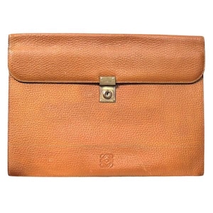May include: A tan leather document portfolio with a pebbled texture, featuring a rectangular flap closure secured by a central brass-toned metal lock mechanism. The lower front section displays a small embossed logo centered on the leather surface.