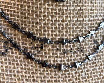 May include: Close-up of multiple layered necklaces with a dark gunmetal finish. The necklaces feature various chain styles, including delicate beaded and star-shaped designs. The jewelry is displayed against a textured, tan burlap background.