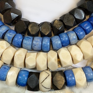 May include: A close-up of a multi-strand necklace made of wooden beads. The necklace features three different colors of beads: black, blue, and white. The beads are all different shapes and sizes, and they are arranged in a random pattern.