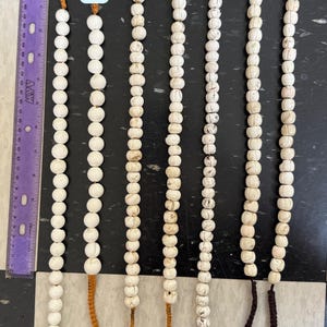 May include: Seven strands of white beaded necklaces with varying cord colors. The beads are round and have a textured appearance. The cords are brown, orange, and tan. A purple ruler is on the left side of the image.