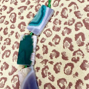 May include: Three rectangular agate pendants in shades of green, blue, and purple are strung together with green thread. The pendants are displayed against a tan background with a brown leopard print pattern.