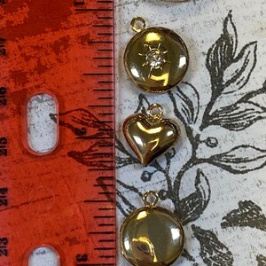 May include: Five gold-tone locket pendants with different designs. The lockets are arranged vertically on a white surface with a red ruler in the background. The lockets feature a round design, a round design with a rhinestone, a heart shape, a round design, and a heart shape.