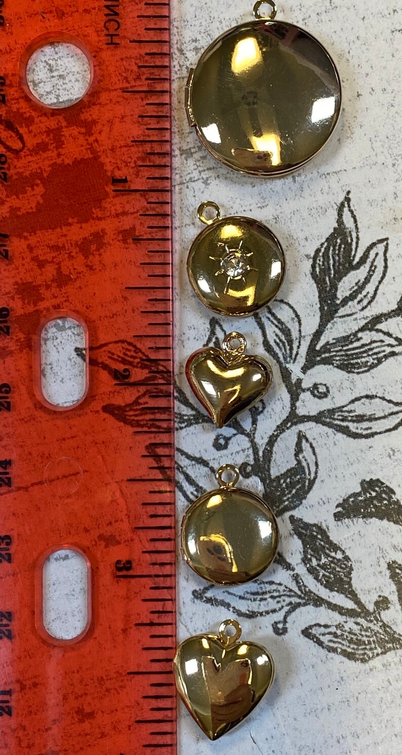 May include: Five gold-tone locket pendants with different designs. The lockets are arranged vertically on a white surface with a red ruler in the background. The lockets feature a round design, a round design with a rhinestone, a heart shape, a round design, and a heart shape.