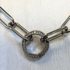 May include: A silver chain necklace with a large, round, silver charm. The charm is encrusted with small, clear stones.