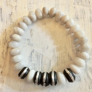 May include: A beaded bracelet featuring a mix of white and gray oval-shaped beads. The bracelet includes a section of black beads with silver stripes. The bracelet is designed to be worn around the wrist.