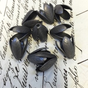 K&ouml;nnte beinhalten: Acht silberne, blumenf&ouml;rmige Perlenkappen aus Metall. Die Kappen haben eine strukturierte Oberfl&auml;che und sind in einem Kreis auf einer wei&szlig;en Oberfl&auml;che mit schwarzer Schrift angeordnet.
