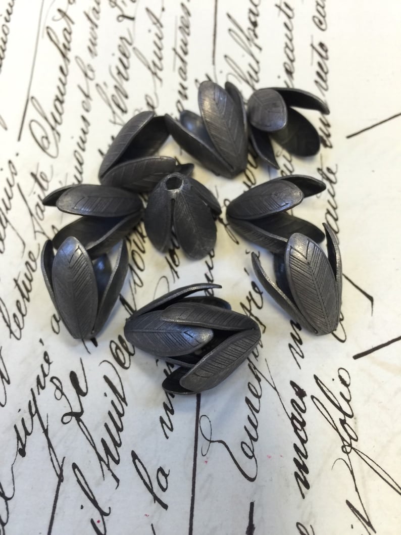 K&ouml;nnte beinhalten: Acht silberne, blumenf&ouml;rmige Perlenkappen aus Metall. Die Kappen haben eine strukturierte Oberfl&auml;che und sind in einem Kreis auf einer wei&szlig;en Oberfl&auml;che mit schwarzer Schrift angeordnet.