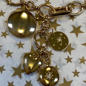 May include: A gold chain necklace with multiple charms, including a heart, a round locket with a star design, and a round locket with a rhinestone. The charms are hanging from a gold chain.