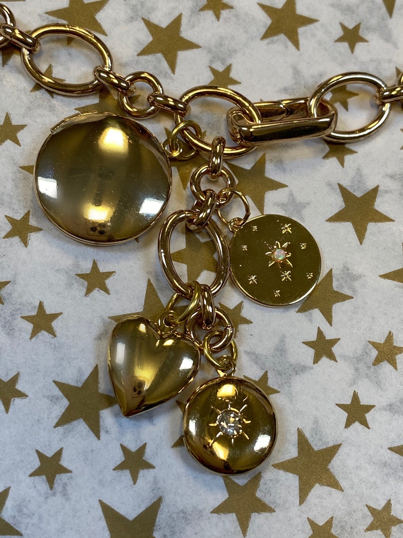 May include: A gold chain necklace with multiple charms, including a heart, a round locket with a star design, and a round locket with a rhinestone. The charms are hanging from a gold chain.