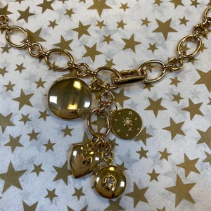 May include: A gold chain necklace with multiple charms, including a heart, a round charm with a star design, and a round charm with a small rhinestone. The necklace is on a white background with gold stars.