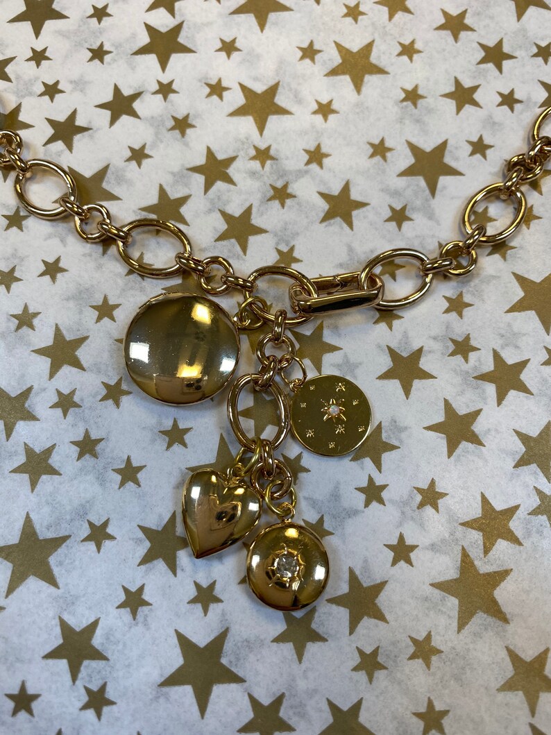 May include: A gold chain necklace with multiple charms, including a heart, a round charm with a star design, and a round charm with a small rhinestone. The necklace is on a white background with gold stars.