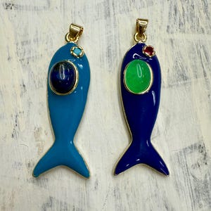 May include: Two fish-shaped pendants with gold-colored trim. One is light blue with a dark blue stone, the other is dark blue with a green stone. Both have gold-colored bails and tail fins.