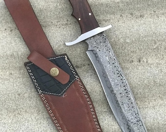 以下が含まれることがあります： 模様入りのダマスカス鋼の刃、3つの金属ピンで固定されたダークブラウンの木製ハンドル、シルバーカラーの鍔を備えた固定刃ナイフ。黒いテクスチャのアクセントが付いたブラウンのレザーシースが付属しています。