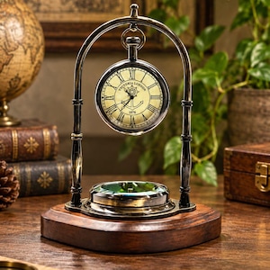Puede incluir: Un reloj de escritorio de latón y madera oscura con esfera circular, números romanos, el texto Victoria London 1876 y un gráfico de ancla. El reloj cuelga de un marco metálico curvo sobre una base de brújula en un soporte de madera.