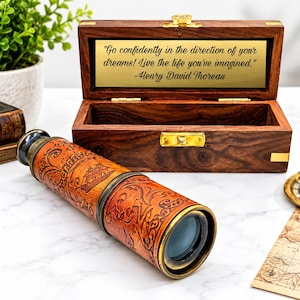 Peut inclure: Une longue-vue nautique en laiton gainée de cuir avec des motifs gaufrés de navires et de volutes, placée devant une boîte en bois ouverte dotée d'une plaque en métal doré gravée de la citation : Go confidently in the direction of your dreams! Live the life you've imagined. ~Henry David Thoreau.