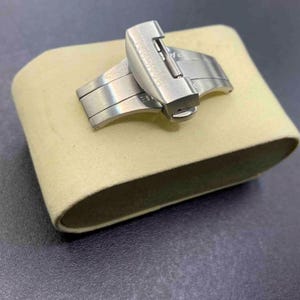 Pode incluir: Um fecho de relógio de metal prateado com acabamento escovado e mecanismo articulado. O fecho está posicionado sobre uma almofada de exibição curva de cor bege claro, destacando a textura metálica e o design funcional da fivela.