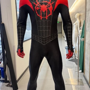 Pode incluir: Um fato de corpo inteiro preto em spandex com um emblema de aranha vermelho no peito e painéis vermelhos nos ombros. O fato tem um padrão de teia de aranha preto texturizado no tronco, braços e pernas, com pontas dos dedos vermelhas nas luvas.
