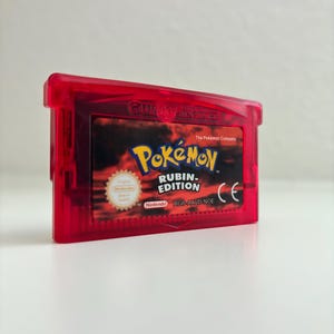 Könnte beinhalten: Ein durchsichtig rotes Game Boy Advance Videospielmodul für Pokémon Rubin Edition. Das Etikett zeigt das Pokémon-Logo, den Text Rubin Edition, das Nintendo-Logo, ein Qualitätssiegel und den Produktcode AGB-AXVD-NOE.