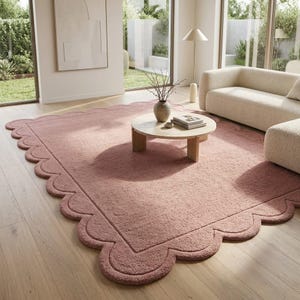 May include: A large, rectangular, dusty rose area rug featuring a distinct scalloped edge border. The rug has a thick, textured pile surface and is positioned on a light-toned wooden floor, extending beneath a round wooden coffee table.