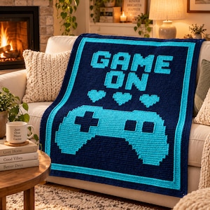 May include: A rectangular crochet blanket featuring a dark navy blue background with a bright cyan border. The center displays the text "GAME ON" in pixelated cyan letters above three cyan pixelated hearts and a large cyan video game controller.