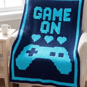 May include: A rectangular crochet blanket featuring a dark navy blue background with a bright cyan border. The design includes the text "GAME ON" in cyan, followed by three cyan heart icons and a large cyan video game controller graphic.