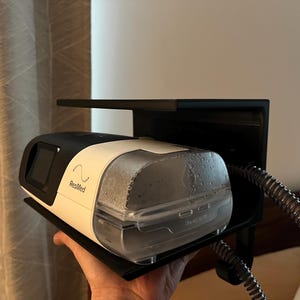 May include: A black and white ResMed CPAP machine with a clear water chamber sits on a black L-shaped shelf mount. The device features a small screen, a side-mounted flexible gray hose, and the ResMed logo printed on the white housing.