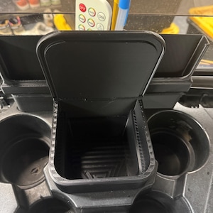 Może przedstawiać: Czarny plastikowy organizer na konsolę środkową z centralnym schowkiem z uchylną pokrywą, otoczony dwoma okrągłymi uchwytami na kubki. Wnętrze schowka posiada trójkątny wzór na dnie, a całość wykonana jest z matowego tworzywa.