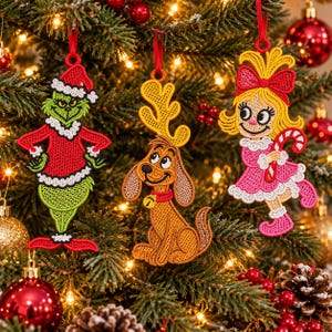May include: Three embroidered Christmas ornaments featuring a green character in a Santa suit, a brown dog with antlers and a bell collar, and a blonde girl in a pink dress holding a candy cane, all hanging from red ribbons on a pine tree.