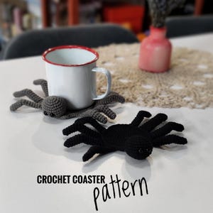 Pode incluir: Dois porta-copos de aranha feitos à mão em crochê, um cinza-escuro e outro preto, cada um com oito pernas e dois olhos escuros. Uma caneca de esmalte branco com borda vermelha repousa sobre o porta-copos cinza. O texto CROCHET COASTER pattern é visível.