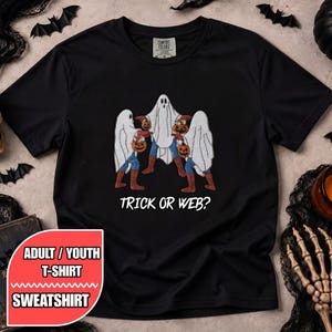 May include: A black short-sleeved t-shirt featuring a graphic of three figures dressed in white ghost costumes over blue and red superhero suits, each holding a jack-o'-lantern. The text below the graphic reads TRICK OR WEB?