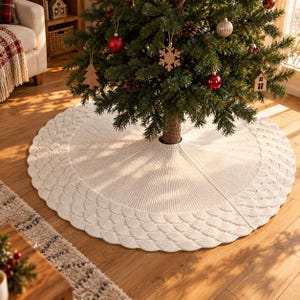 Könnte beinhalten: Ein kreisförmiger, cremeweißer gestrickter Weihnachtsbaumteppich mit einem strukturierten, bogenförmigen Randmuster. Der Stoff weist ein geripptes Design auf, das von der mittleren Öffnung ausgeht und eine neutrale Basis für den Baum bietet.