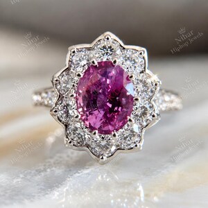 Könnte beinhalten: Ein silberfarbener Ring mit einem zentralen, oval geschliffenen, leuchtend rosa Edelstein, umgeben von einem Kranz aus zwölf runden, klaren Steinen in Blütenform. Die Ringschiene ist an den sichtbaren Seiten mit kleinen klaren Steinen besetzt.