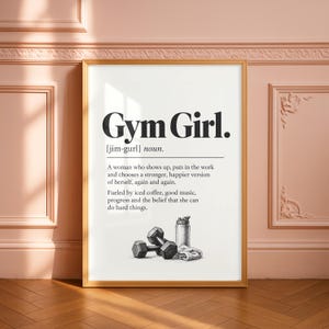 Pode incluir: Uma impressão de arte de parede emoldurada com o texto Gym Girl, definido como uma pessoa que aparece e faz o trabalho. Abaixo da definição, uma ilustração em preto e branco mostra um par de halteres, uma garrafa de água e uma toalha.