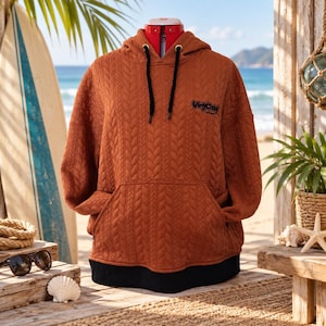Peut inclure: Un sweat à capuche couleur rouille avec une texture matelassée en tricot torsadé, une capuche à cordons noirs et un ourlet noir. La poitrine porte le texte brodé en noir "VinyCra" avec un texte plus petit en dessous sur un mannequin.