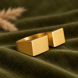 Op de afbeelding: Een goudkleurige open ring met twee rechthoekige, platte uiteinden met een geborstelde, matte afwerking. De band is cirkelvormig en de twee vierkante uiteinden zijn bij de opening geplaatst, wat zorgt voor een geometrisch ontwerp.