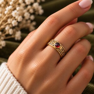 May include: A gold-toned band ring featuring an intricate embossed Celtic knot pattern encircling the entire circumference. The center of the ring is set with a single, smooth, round, deep red gemstone that sits flush within the metal band.