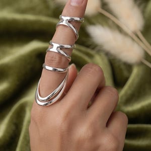 Puede incluir: Un conjunto de anillos de tono plateado usados en un solo dedo, con un diseño de múltiples bandas. La joyería consta de dos anillos superiores con estructura cruzada y un anillo más grande, liso y de doble bucle en la base, creando un aspecto en capas.